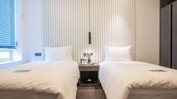 A hotel room with two beds, a bedside table with a phone and a lamp, and a window with blinds.