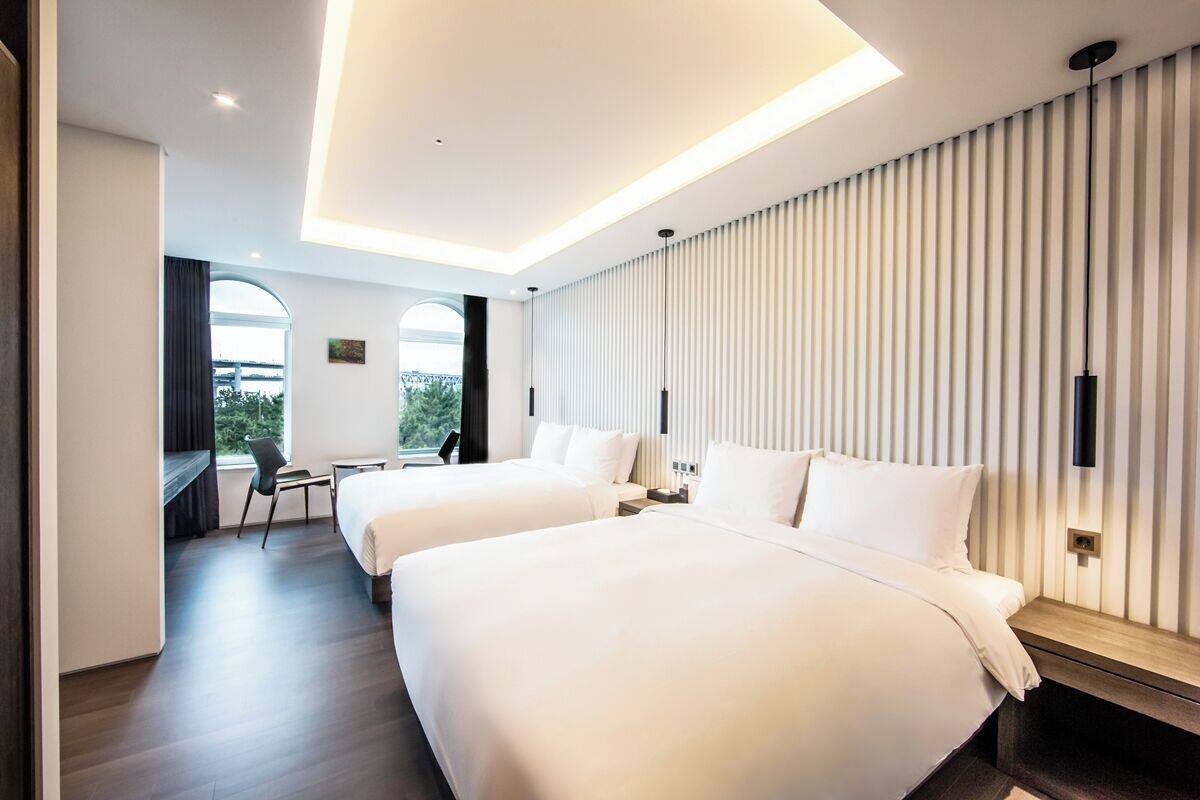 A modern hotel room with two beds, a desk, and a view of greenery through large windows.