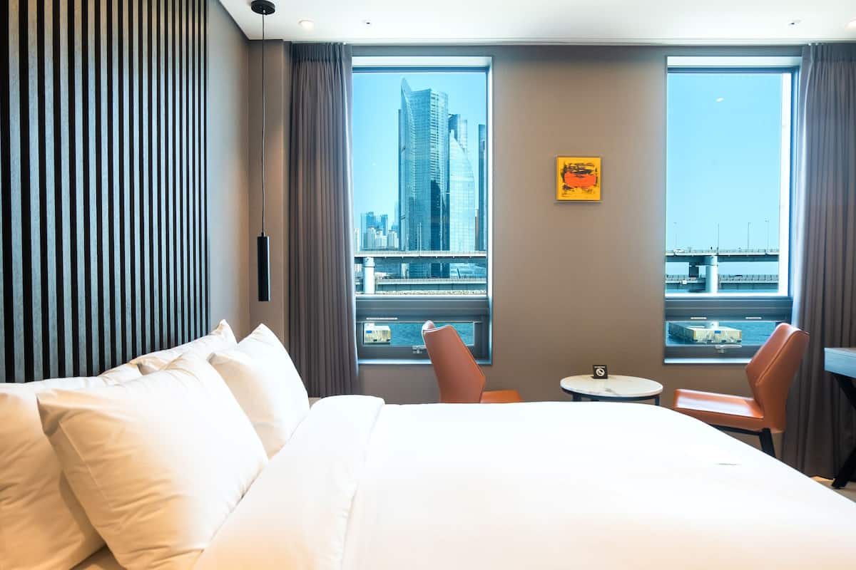 A hotel room with a large bed, two chairs, a small table, and a view of a cityscape through large windows.