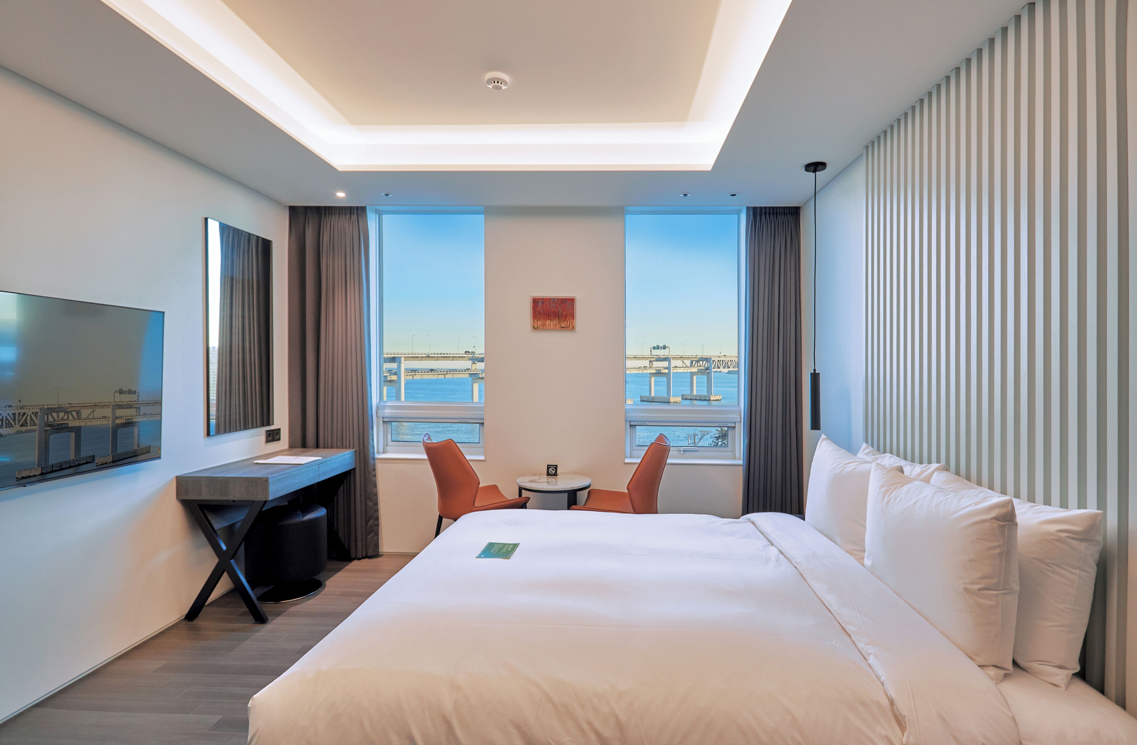 A modern hotel room with a large bed, a desk, a TV, and a view of the cityscape.
