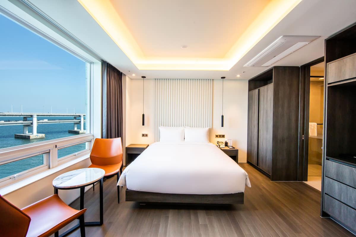 A modern hotel room with a large bed, a small table, two chairs, a TV, and a view of the water through large windows.