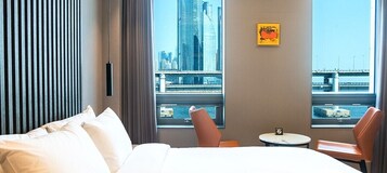 A hotel room with a bed, a small table, two chairs, a window with a city view, and a painting on the wall.