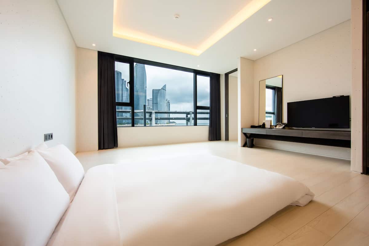 A modern hotel room with a large bed, a TV, and a city view through large windows.