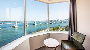 A room with a large bay window overlooking a bridge and water. There is a small round table and a chair with a logo. - AG405 Hotel (Busan)