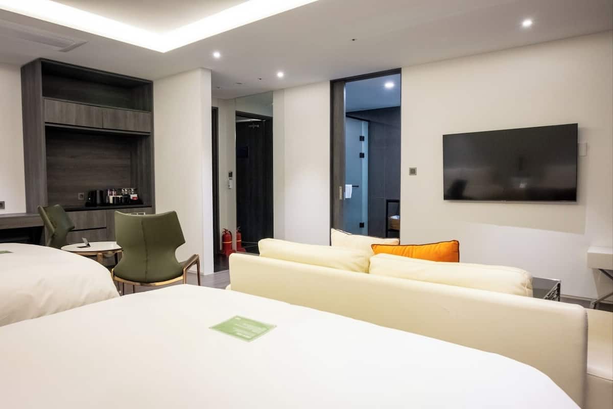 A modern hotel room with a large bed, a sofa, a desk with a chair, and a flat-screen TV mounted on the wall.