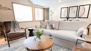 A modern living room with a sectional sofa, a leather armchair, a round wooden coffee table, and a large window providing natural light. - Peaceful modern oasis -walking distance to Village (Sunriver)
