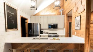 A modern kitchen with wooden walls, a white countertop, stainless steel appliances, and three bar stools. - Peaceful modern oasis -walking distance to Village (Sunriver)