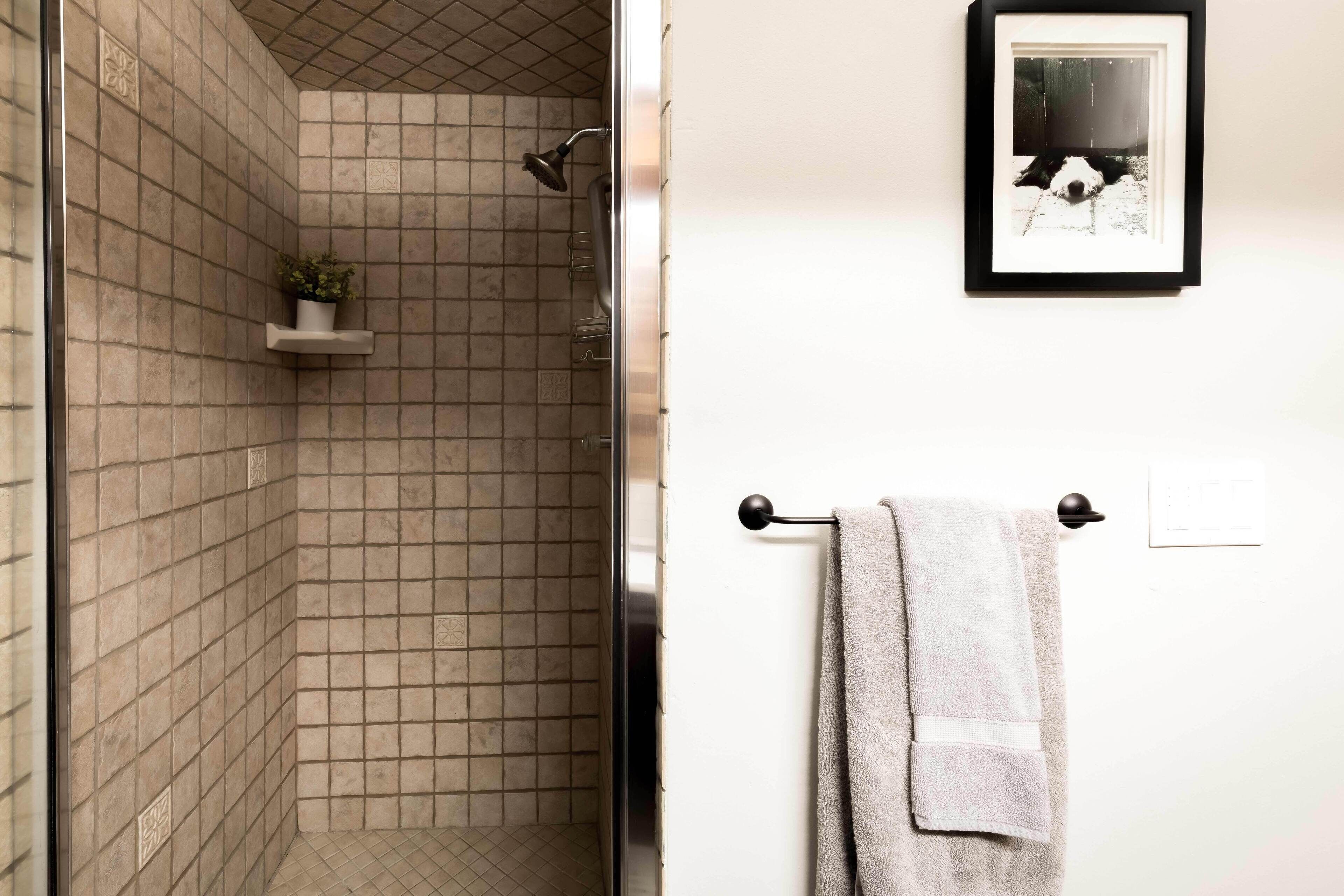 A bathroom with beige tiled walls, a shower, a towel rack with a towel, and a framed picture on the wall.