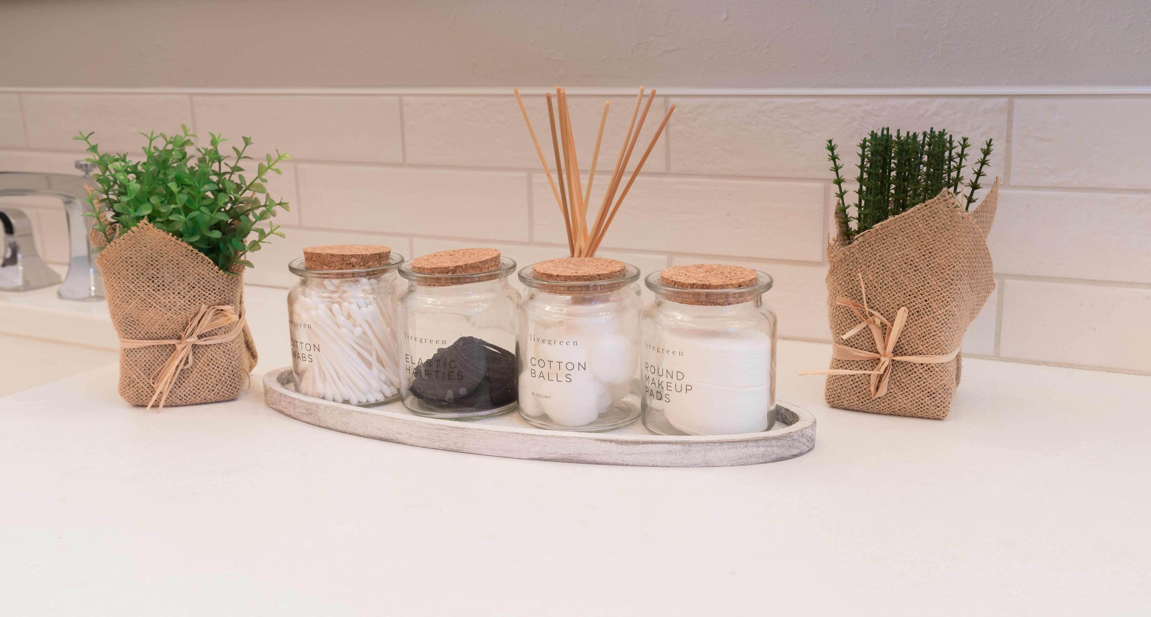 A kitchen counter with jars of cotton balls, elastic hair ties, and round makeup pads, each with a cork lid and tied with twine.