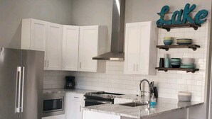 Una cocina moderna con armarios blancos, una campana extractora de acero inoxidable, un horno empotrado, un microondas y una cafetera. Hay taburetes altos azules y una repisa con vajilla.