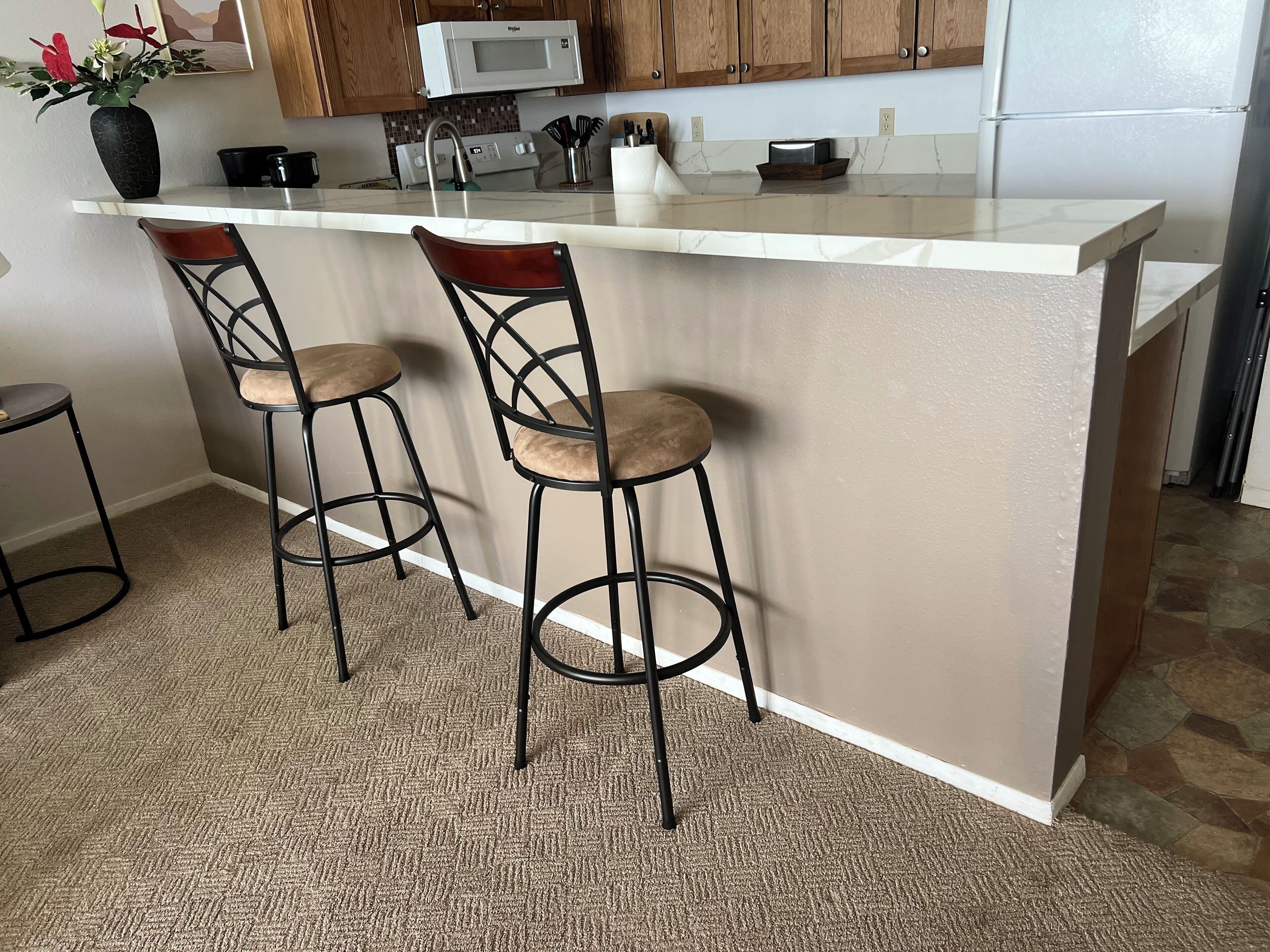 A kitchen with a white marble countertop, two bar stools, a microwave, and wooden cabinets.