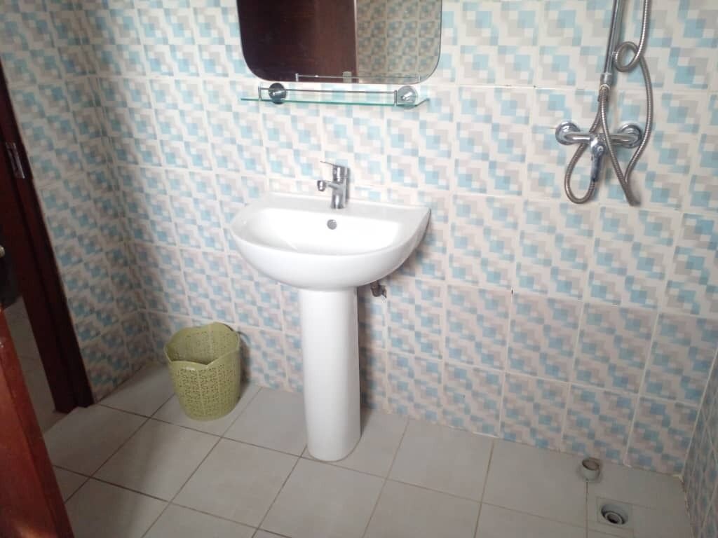 A bathroom with a white pedestal sink, a glass shelf above the sink, a shower with a handheld showerhead, and a green trash bin.