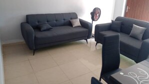 A living room with two dark sofas, a standing fan, and a wooden door in the background. - Lovely 1-Bed duplex house (BINGERVILLE)