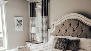 A neatly made bed with a tufted headboard, a bedside table with a lamp, and a window with patterned curtains. - Robin’s Nest Cabin- quiet cove on Lake Hamilton (Hot Springs National Park)