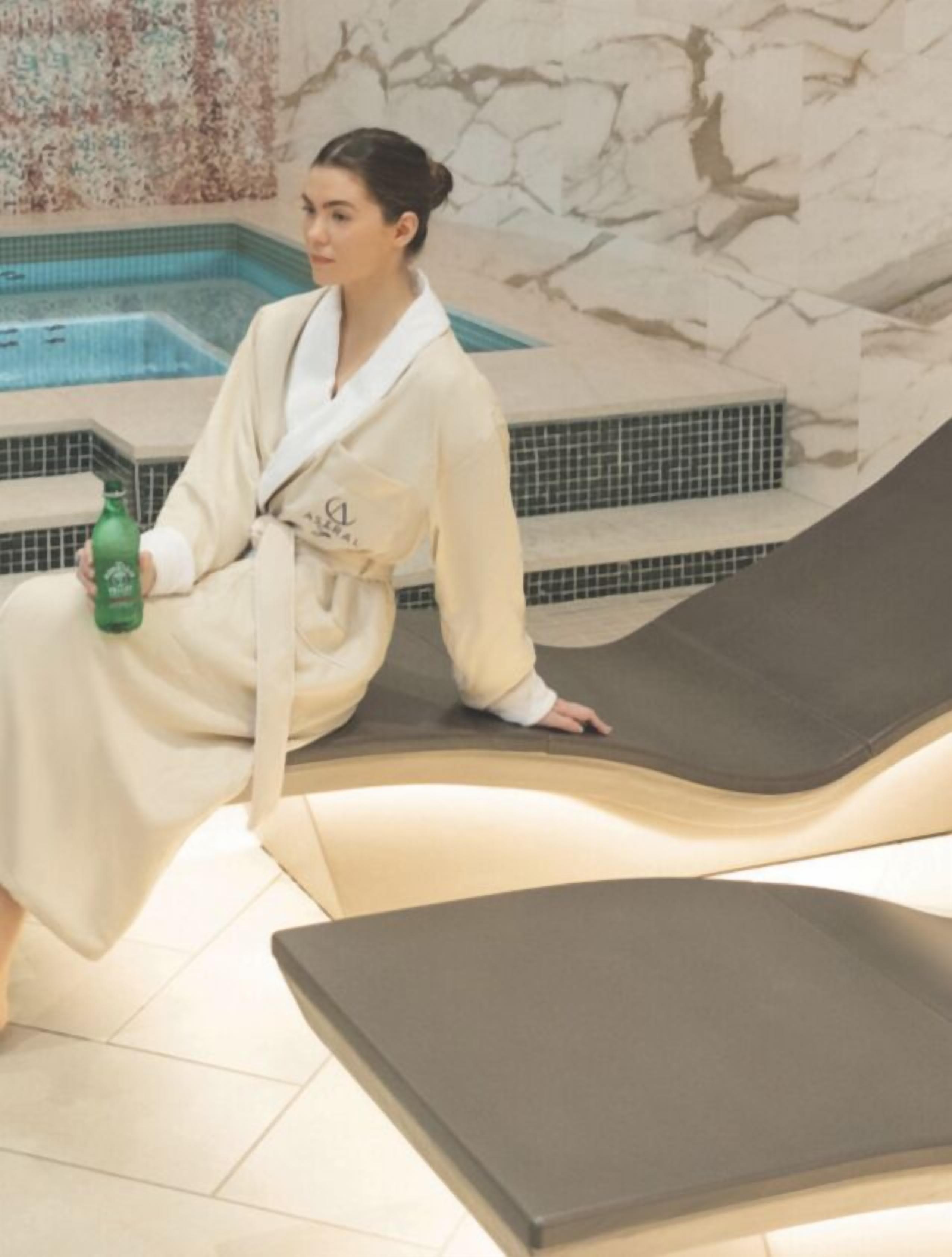 Ein Wellnessbereich mit Marmorwand, einem Pool und einer Person im Bademantel, die eine Flasche hält.