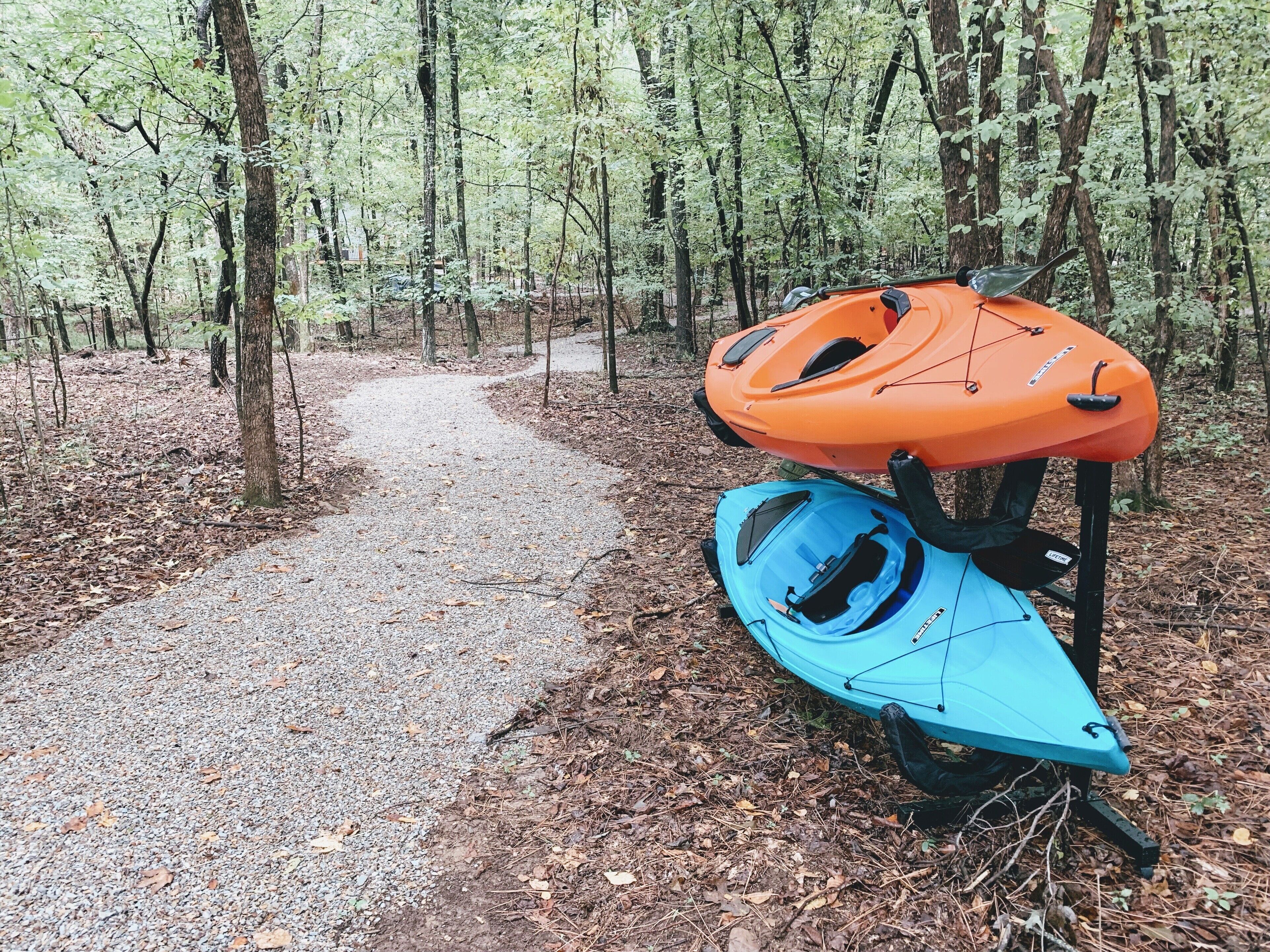 Kayaks and a paddleboard on a gravel path in a forest.