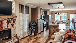 A spacious living room with a fireplace, a ceiling fan, and a large TV mounted on a stone wall. - Robin’s Nest Cabin- quiet cove on Lake Hamilton (Hot Springs National Park)