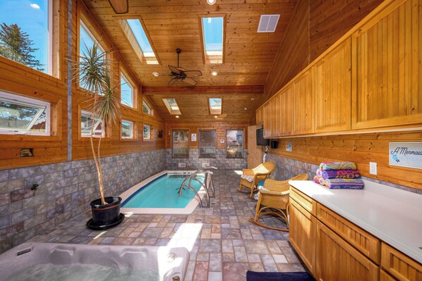 An indoor swimming pool with wooden walls and ceiling, stone tiles, and a seating area with chairs.