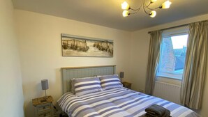 A bedroom with a bed, a nightstand, a lamp, a window with curtains, and a painting on the wall. - 2 Bedroom Riverside Penhouse Apartment St Neots (Saint Neots)