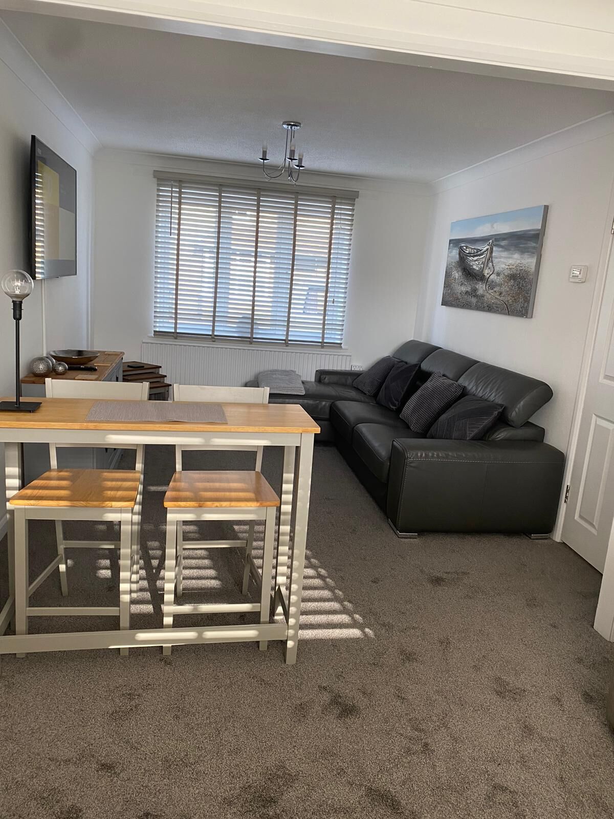 A modern living room with a black leather sofa, a wooden dining table with two chairs, a chandelier, and a large window with blinds.