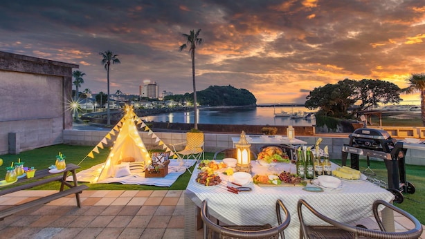 A picnic setup with a table, chairs, and a teepee tent on a patio overlooking a body of water and cityscape at sunset. - West coast villa Shirahama (Shirahama)