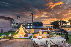 A picnic setup with a table, chairs, and a teepee tent on a patio overlooking a body of water and cityscape at sunset. - West coast villa Shirahama (Shirahama)