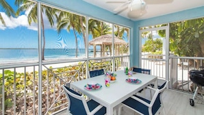 A dining area with a table set for two, large windows with a view of palm trees and a beach, and a ceiling fan. - Kaibo Kai Hacienda B-5 2BR/2BA at Scenic Kaibo Kai -- Beach & Pool (Cayman Kai)