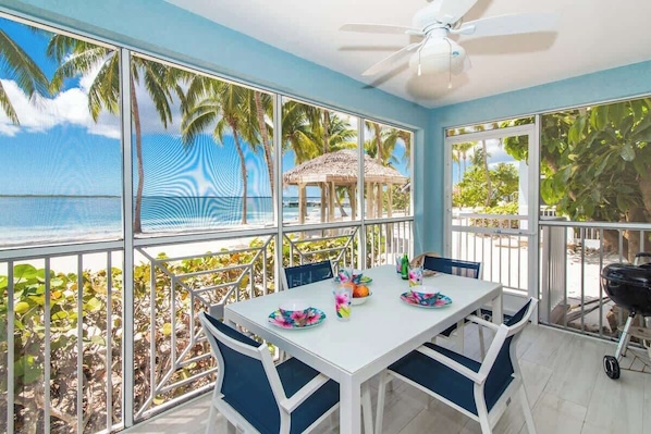 A dining area with a table set for two, large windows with a view of palm trees and a beach, and a ceiling fan.