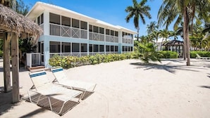 A beachfront hotel with a light blue facade, multiple balconies, and a sandy beach area with lounge chairs. - Kaibo Kai Hacienda B-5 2BR/2BA at Scenic Kaibo Kai -- Beach & Pool (Cayman Kai)