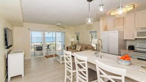 A modern kitchen with white cabinetry, a dining area with white chairs, and a large window overlooking a balcony. - Kaibo Kai Hacienda B-5 2BR/2BA at Scenic Kaibo Kai -- Beach & Pool (Cayman Kai)