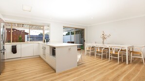 A modern kitchen with white cabinetry, a central island, and a dining area with white chairs and a table. - Meeseeks - Pet Friendly - 2 Mins Walk to Beach (Culburra Beach)
