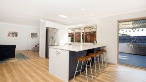A modern kitchen with a central island, bar stools, and a view of a patio with a grill. - Meeseeks - Pet Friendly - 2 Mins Walk to Beach (Culburra Beach)