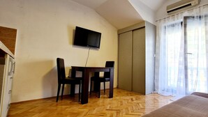 Een kamer met een houten visgraatvloer, een bed met een beddegoed met dessin, een aan de muur bevestigde televisie, een tafeltje met twee stoelen en een schuifdeur met gordijn met patroon.
