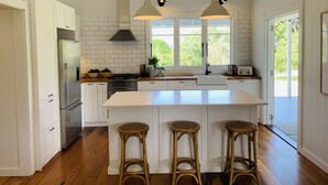 A modern kitchen with white cabinetry, a stainless steel refrigerator, a gas stove, and a large island with bar stools. - Meryta House (Norfolk Island)