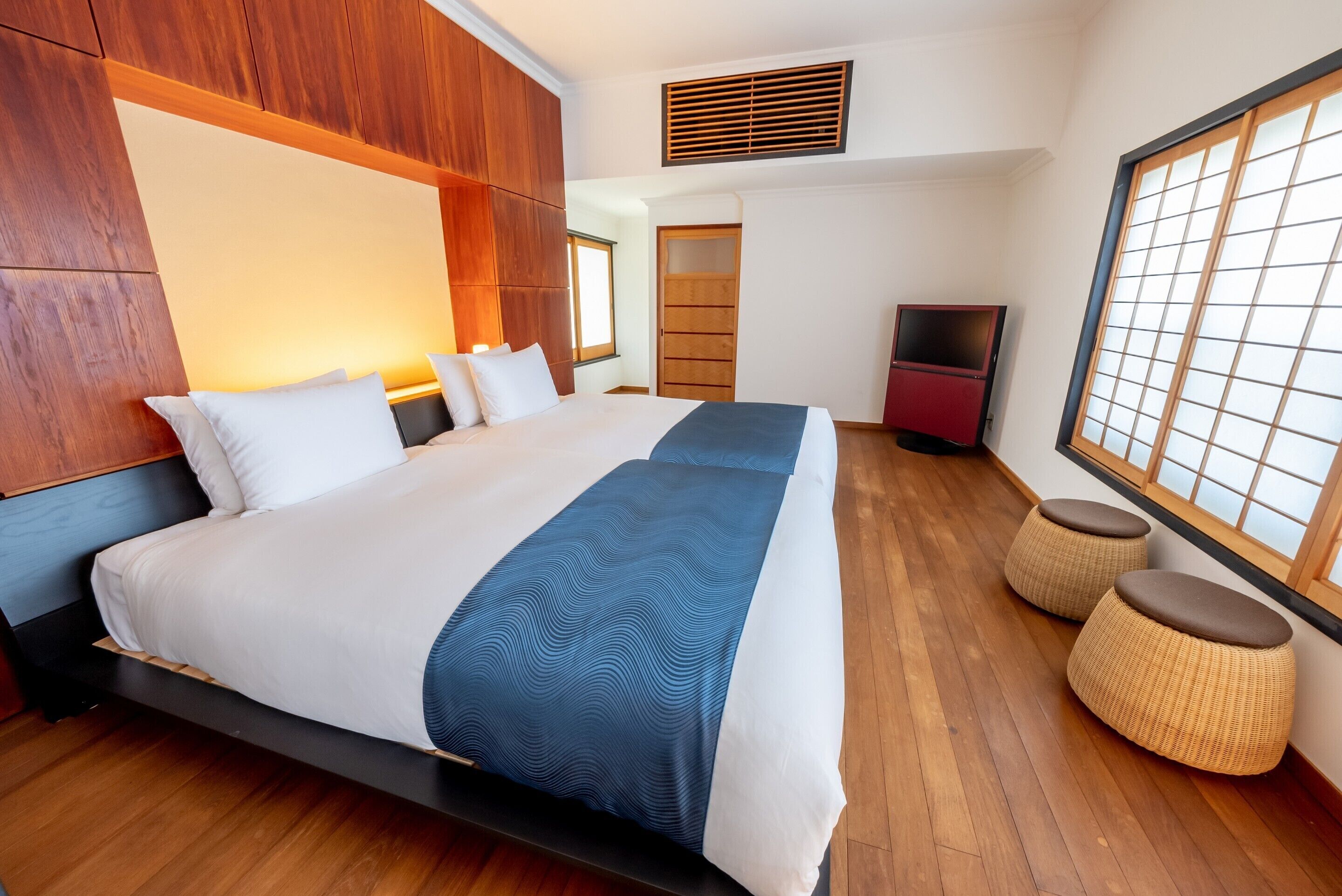 Una habitación de hotel moderna con una cama grande, paneles de madera, un televisor y vistas a través de un celosía japonesa.