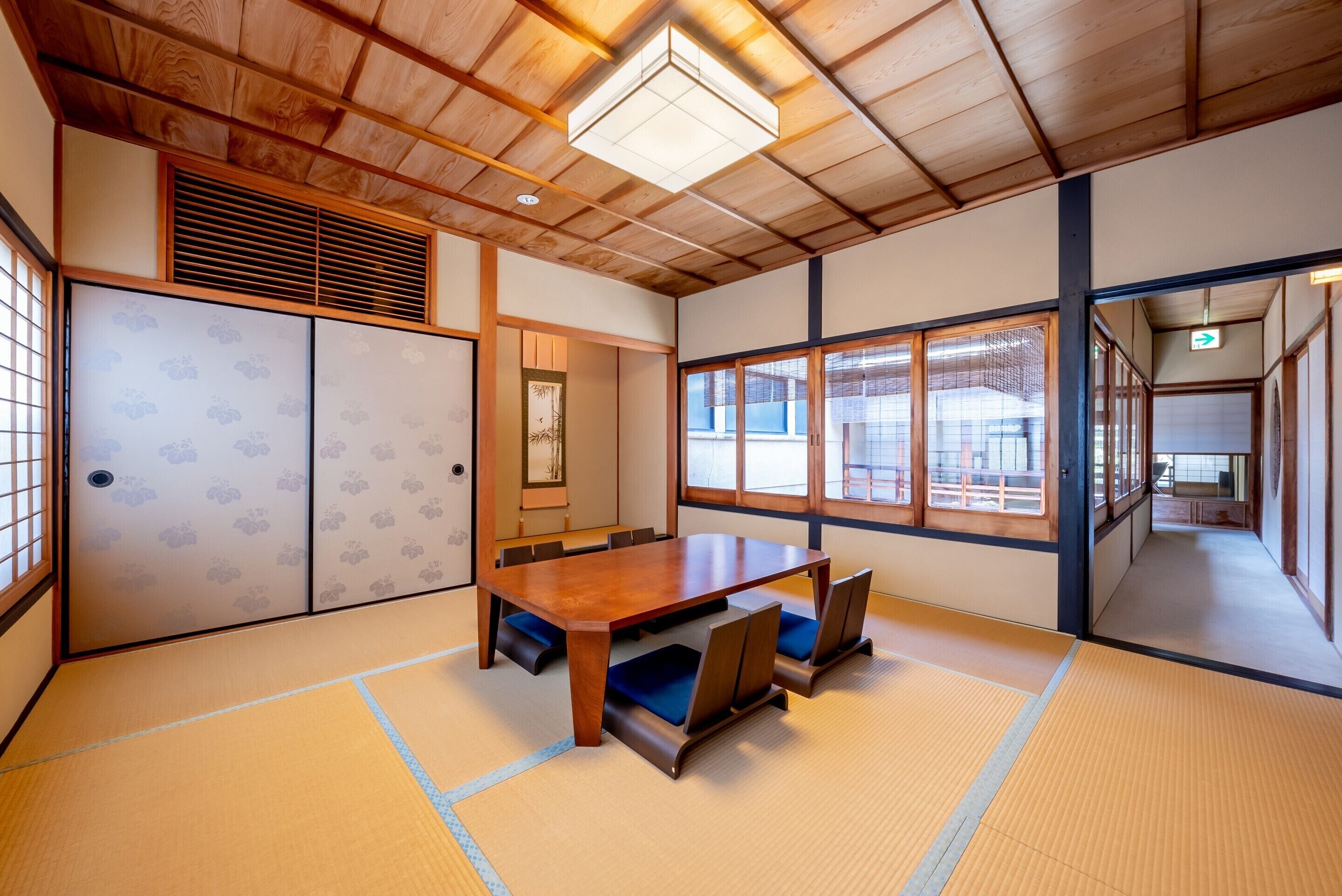 A traditional Japanese room with tatami flooring, a wooden table with cushions, sliding doors, and large windows.