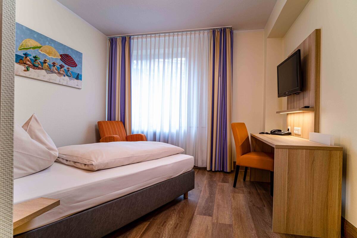 A hotel room with a bed, a desk, a chair, a television, and a painting on the wall.