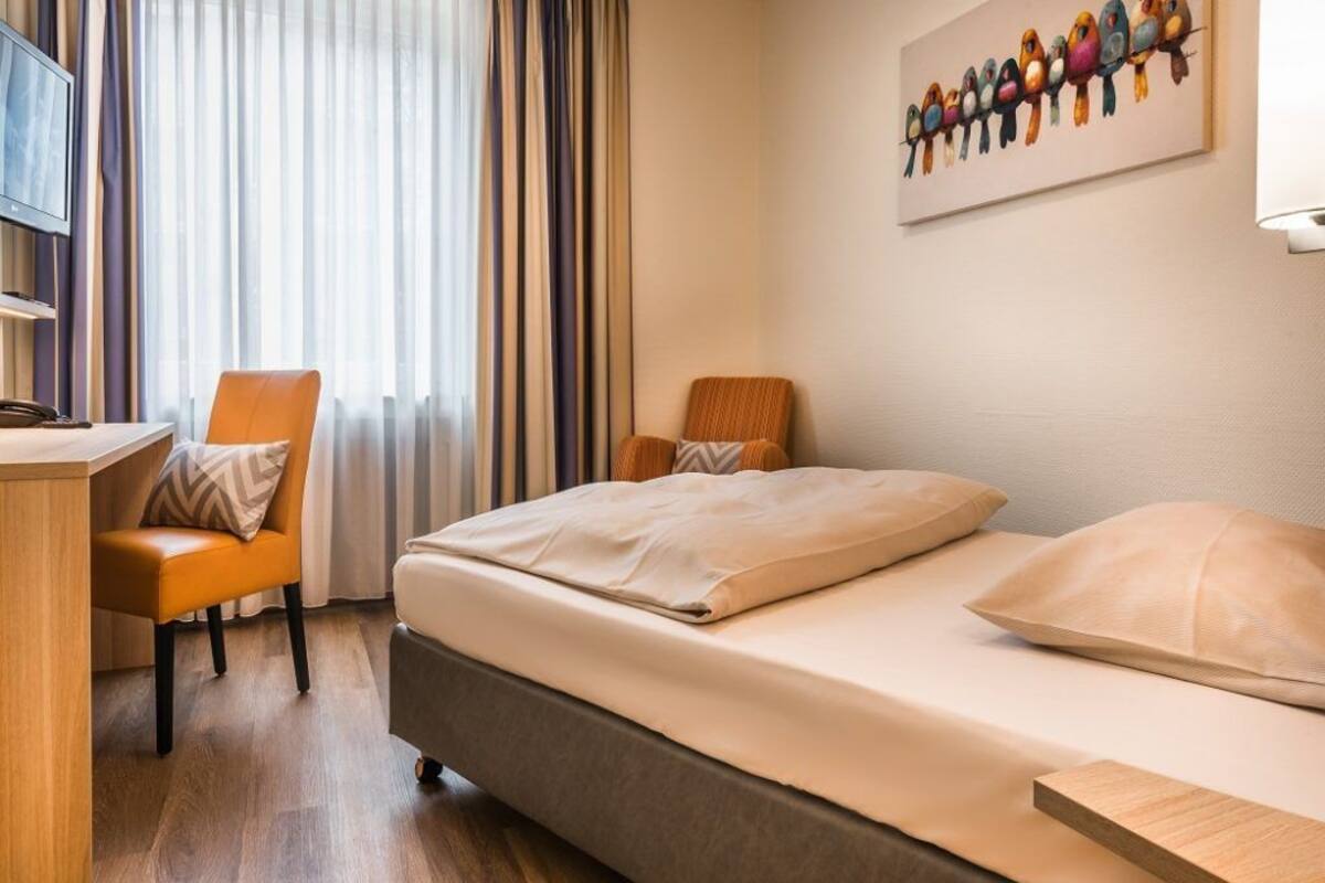 A hotel room with a bed, two chairs, a desk, and a painting on the wall.