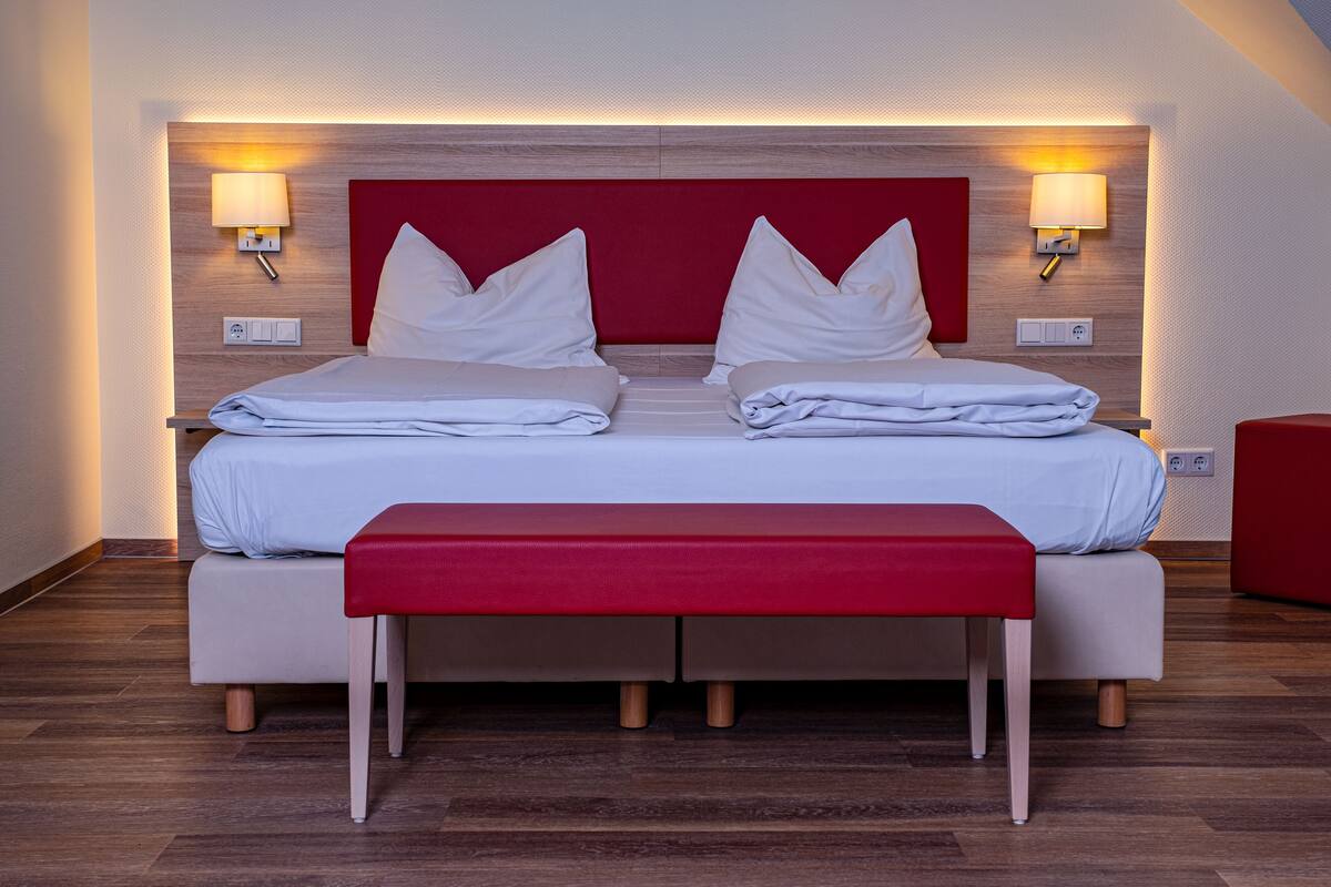 A double bed with a red headboard, two bedside tables with lamps, a red bench, and wooden flooring.