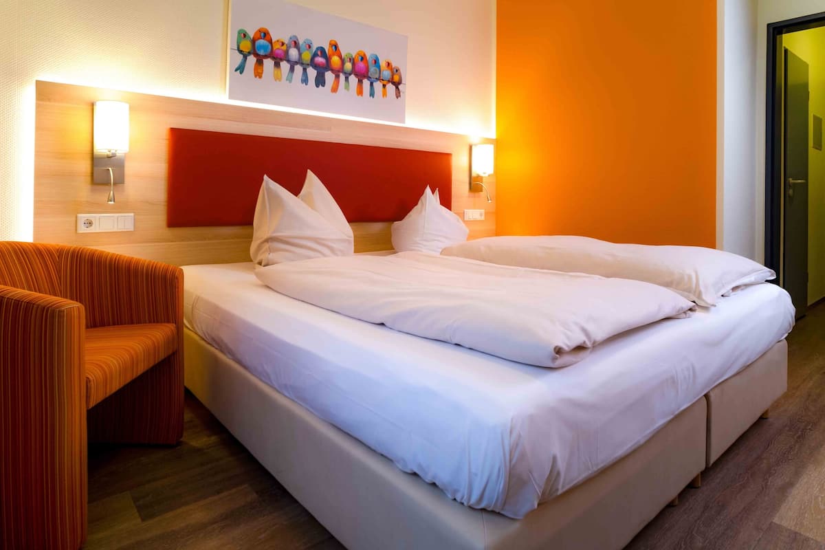 A hotel room with a large bed, a chair, a wall-mounted light, and a painting of birds.