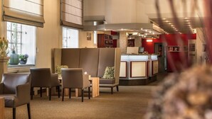 A hotel lobby with seating areas, a reception desk, and a view of the outside through large windows. - Hotel am Stadtpark (Hilden)