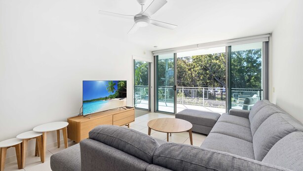 A modern living room with a grey sofa, a TV on a wooden cabinet, and a balcony with a glass door.