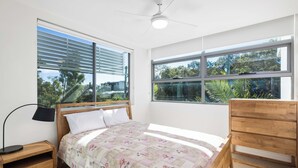 A bedroom with a wooden bed, a nightstand, a desk, and a window with blinds.