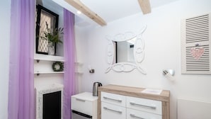 A modern bathroom with a white vanity, a mirror, a small shelf with a potted plant, and a window with purple curtains. - Lavender Room (Dubrovnik)