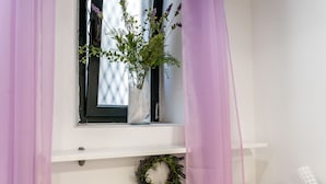 A window with a white vase containing flowers, a wreath on the lower shelf, and a wall-mounted light fixture. - Lavender Room (Dubrovnik)