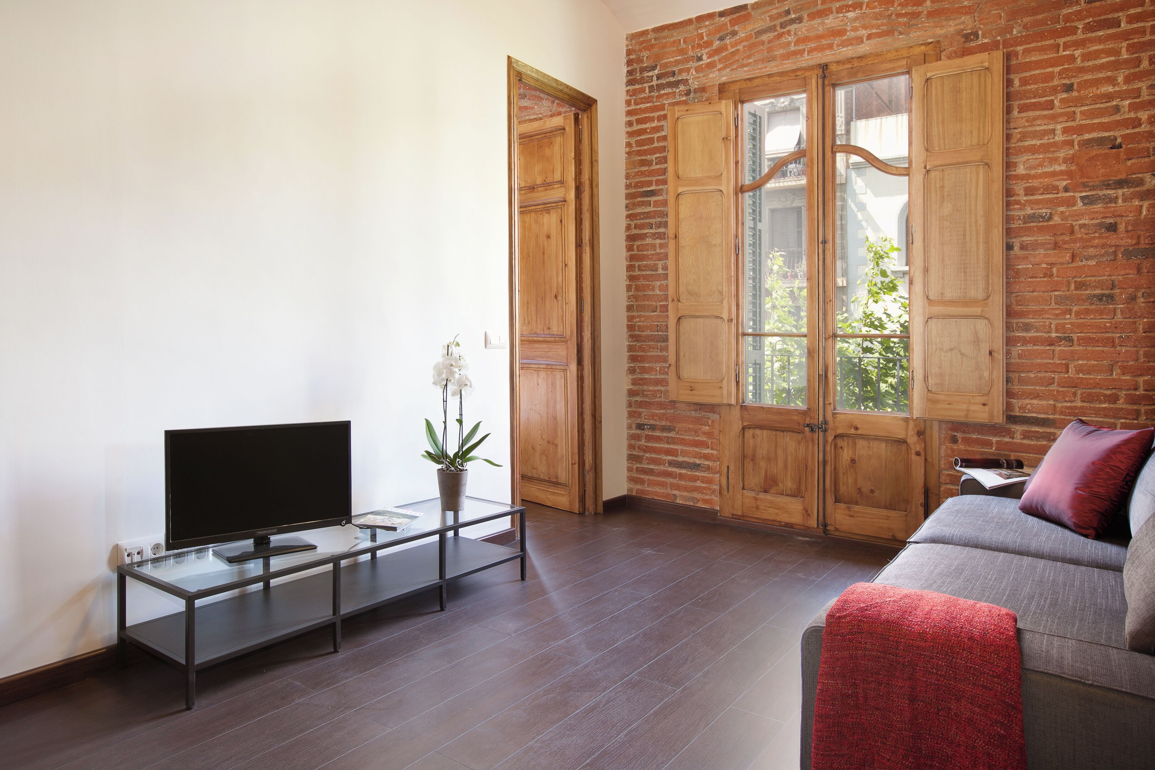 A modern living room with a flat-screen TV on a glass table, a grey sofa with a red throw blanket, and wooden doors with glass panels.