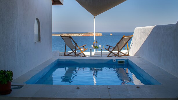 Ein Poolbereich mit zwei Liegestühlen und einem Tisch, mit Blick auf ein Gewässer und eine felsige Küste.