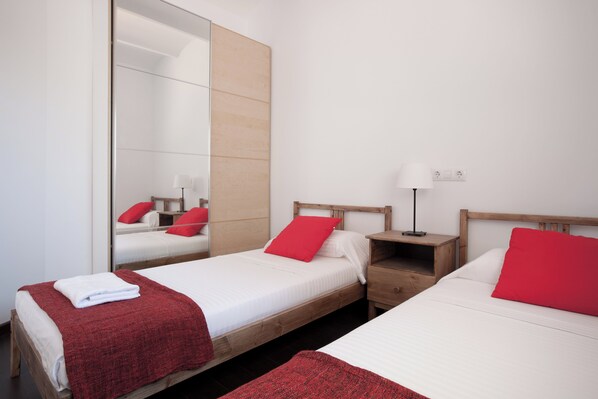 A hotel room with two single beds, each with a red pillow and a white blanket. There is a wooden nightstand with a lamp between the beds. A large mirror is mounted on the wall above the left bed.