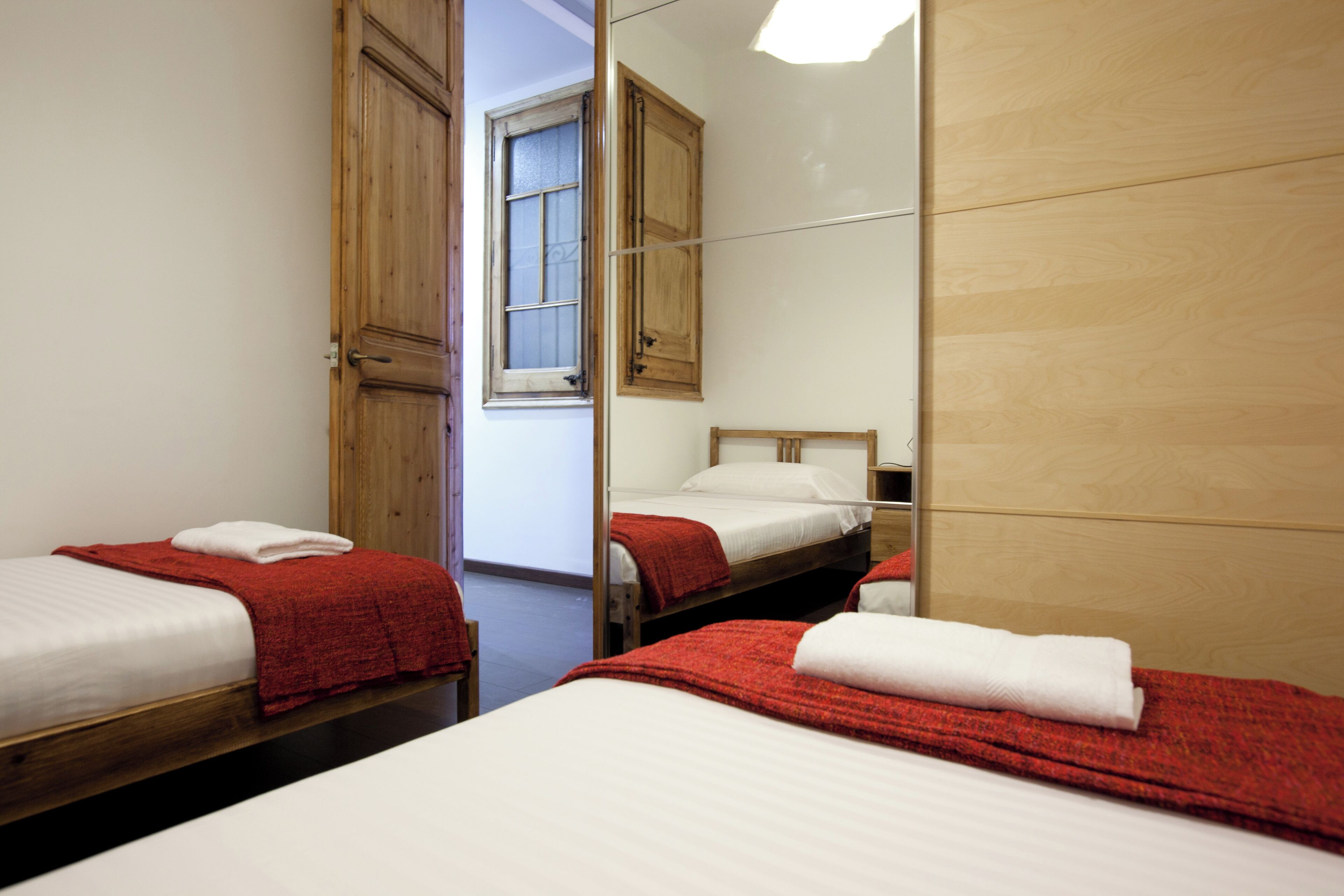 Habitación de hotel con dos camas, cada una con una manta roja y almohada blanca, una puerta de madera y ventana con vistas.