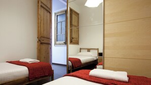 Habitación de hotel con dos camas, cada una con una manta roja y almohada blanca, una puerta de madera y ventana con vistas.